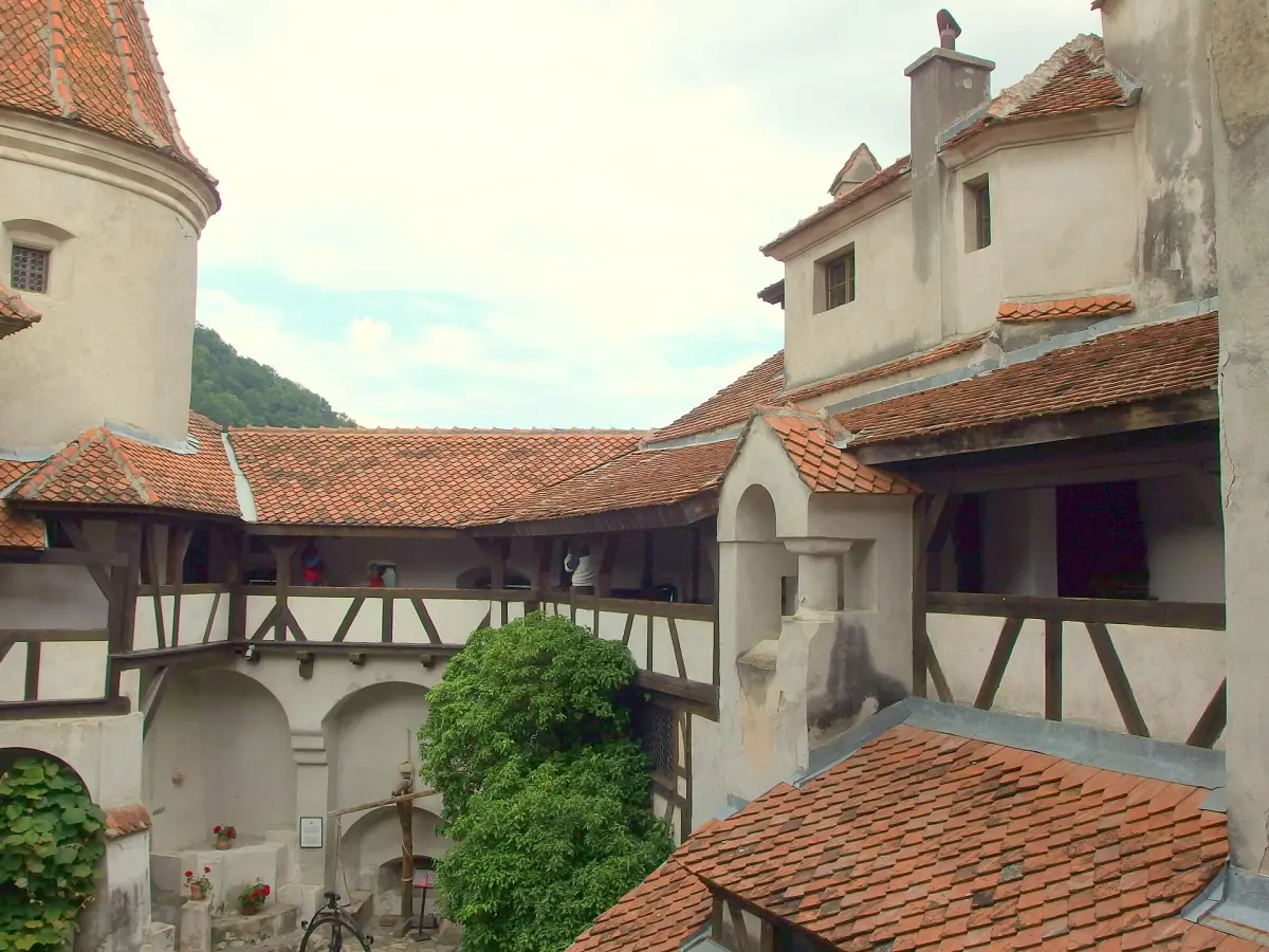 Bran Castle courtyard with red tiled roofs, a round tower and a green tree below a cloudy sky