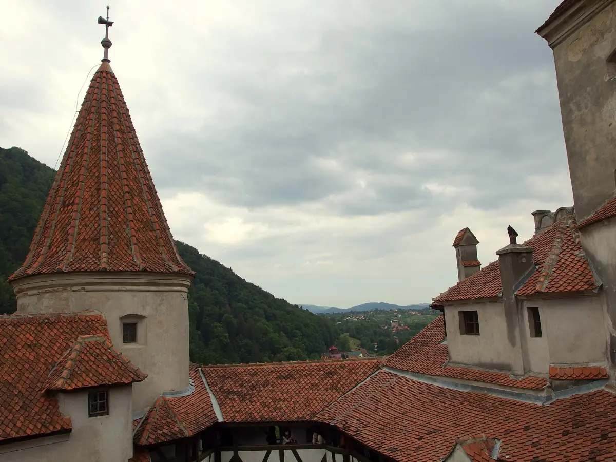 Conical tower roof and courtyard roofs with forested hills behind under cloud