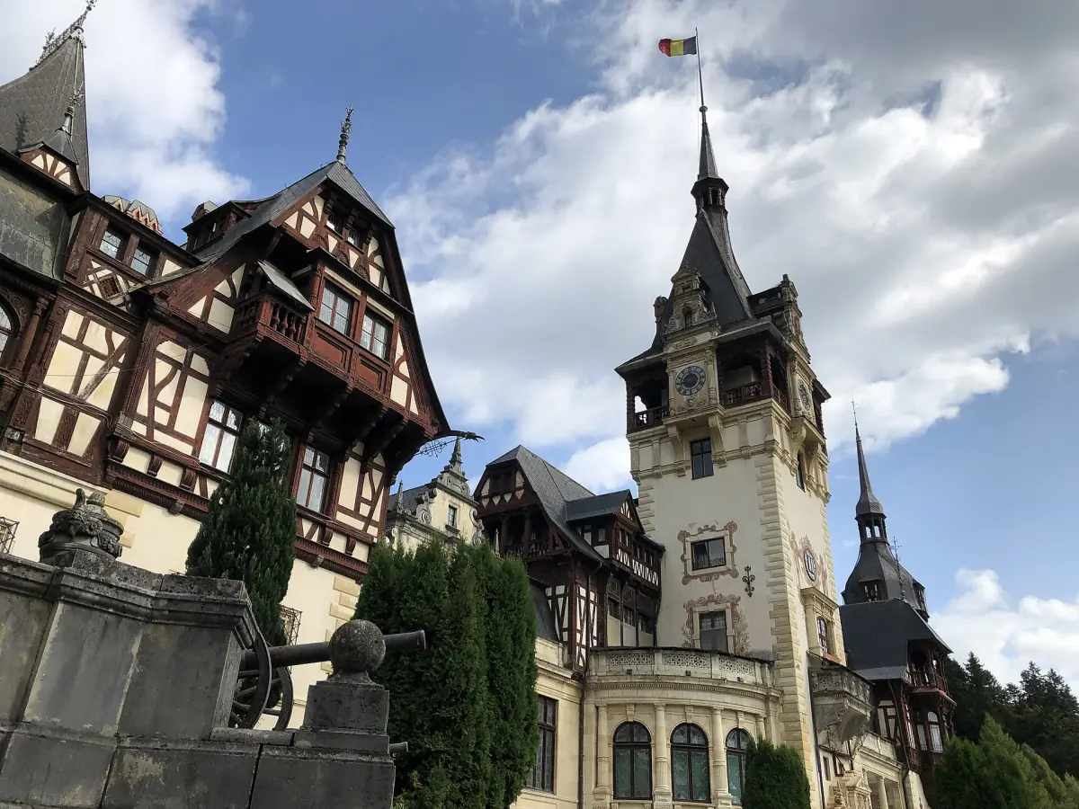 Half timbered upper floors and the tall clock tower of Peleș Castle under a sky with white clouds
